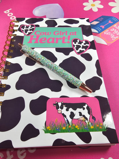 Notebook with cow print design, pen, and decorative elements on a pink background