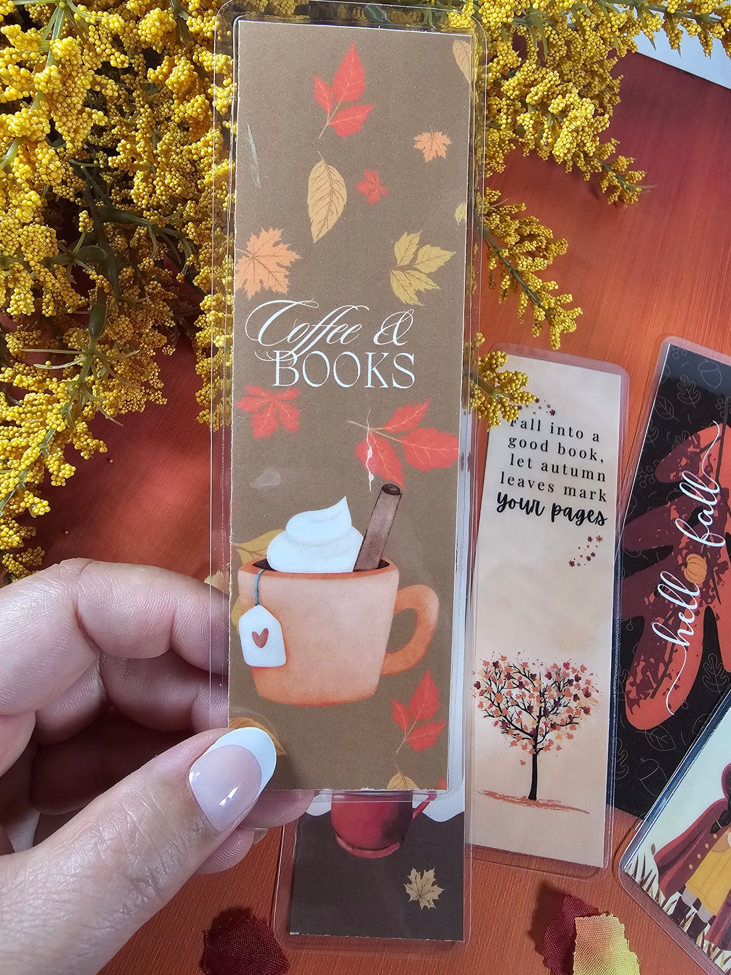 Decorative bookmarks with autumn-themed designs held by a hand, surrounded by yellow flowers and more bookmarks.