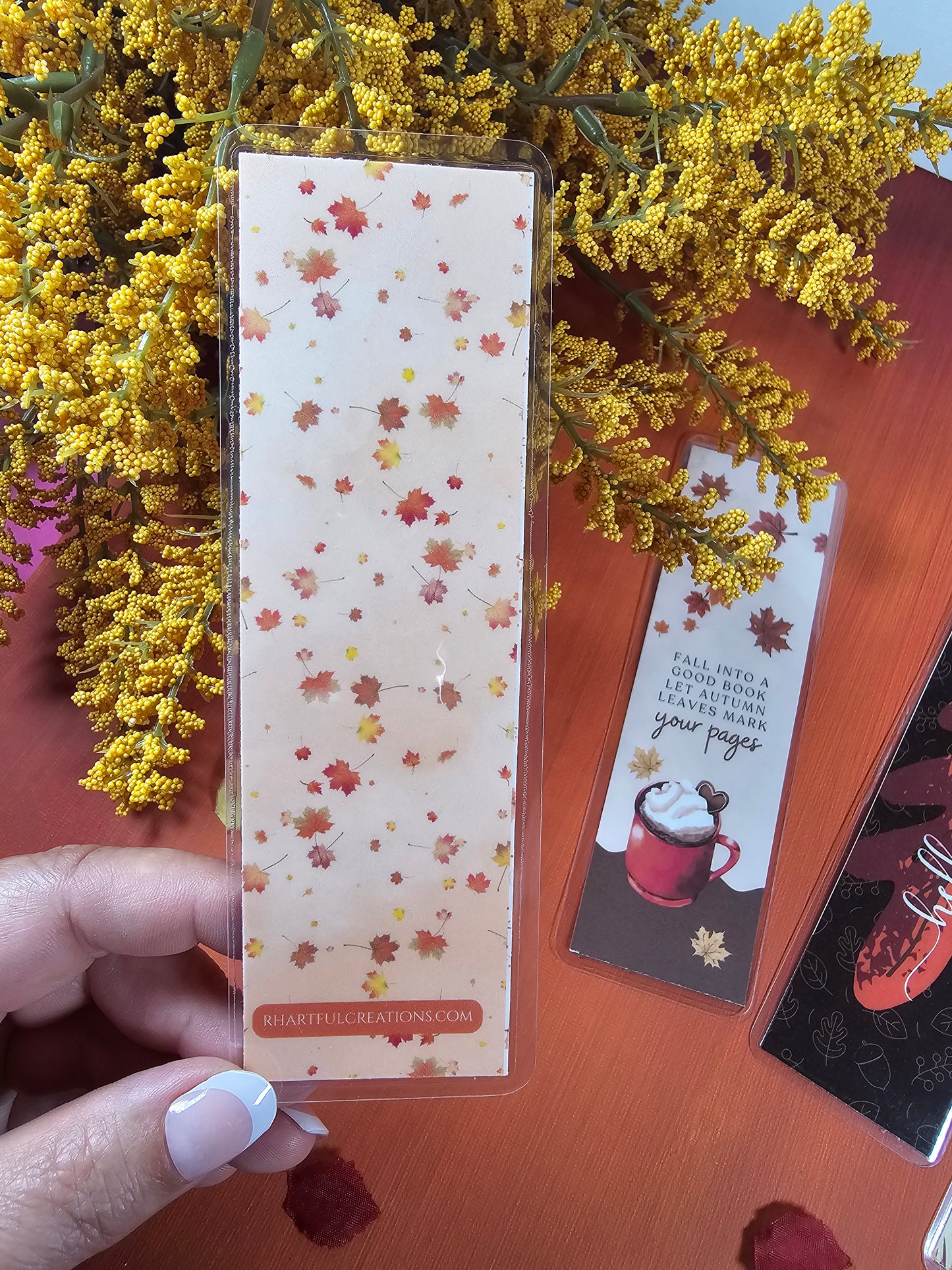 Decorative paper with autumn leaf pattern held by a hand, surrounded by yellow flowers on a wooden surface.