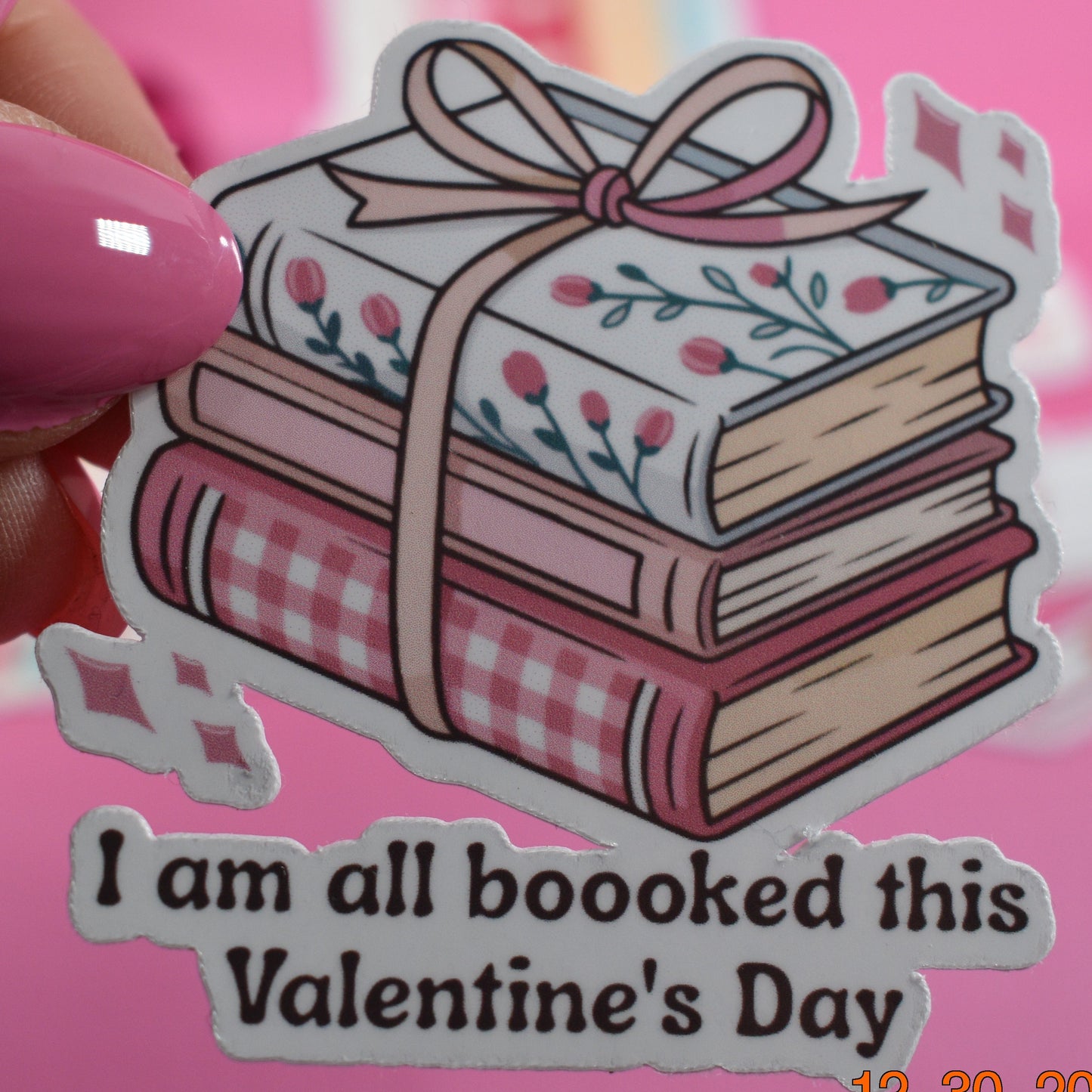Sticker of books with a bow and text 'I am all booked this Valentine's Day' held by a hand with pink nail polish on a pink background.