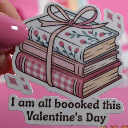 Sticker of books with a bow and text 'I am all booked this Valentine's Day' held by a hand with pink nail polish on a pink background.