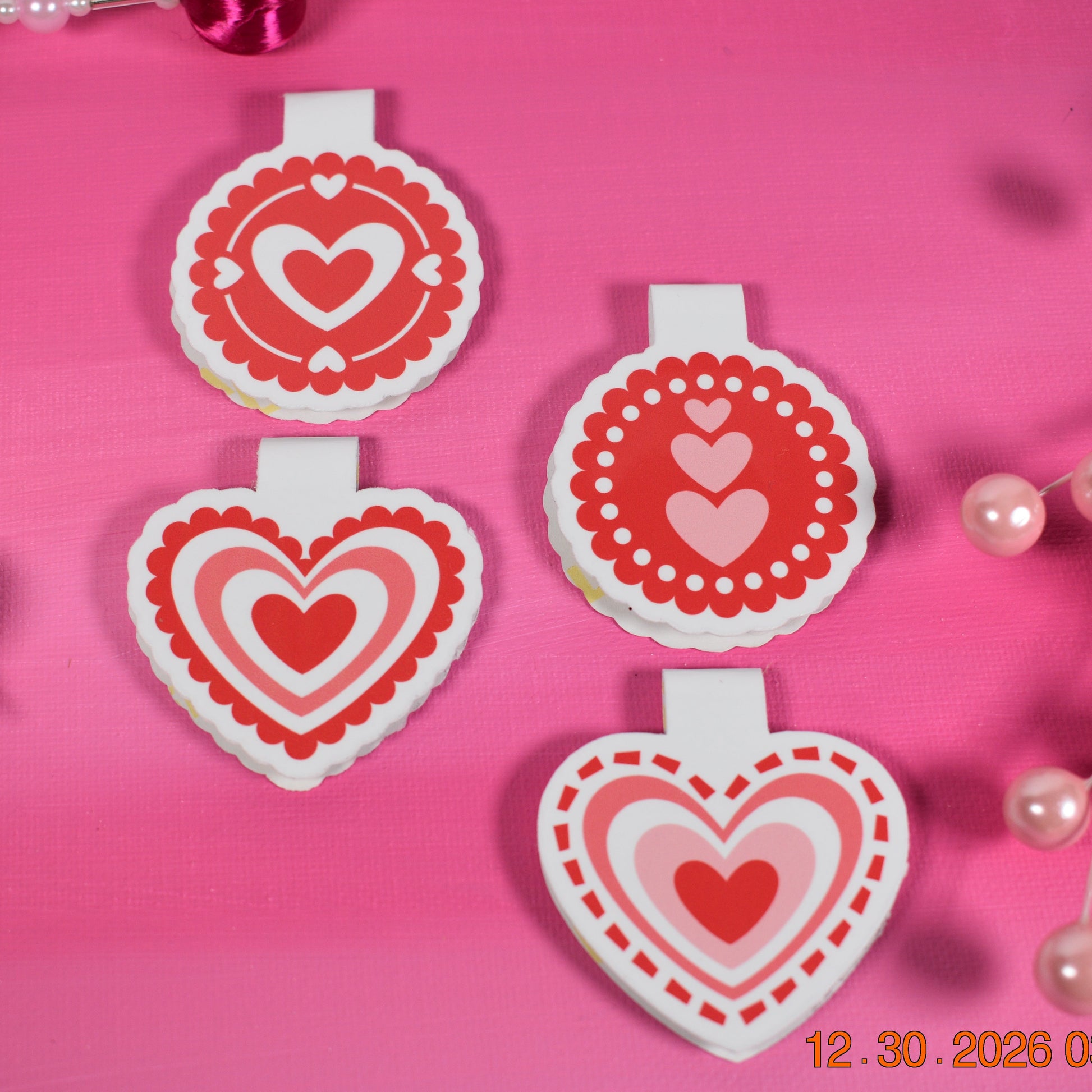 Four Valentine heart magnetic bookmarks on a bright pink background with decorative beads.