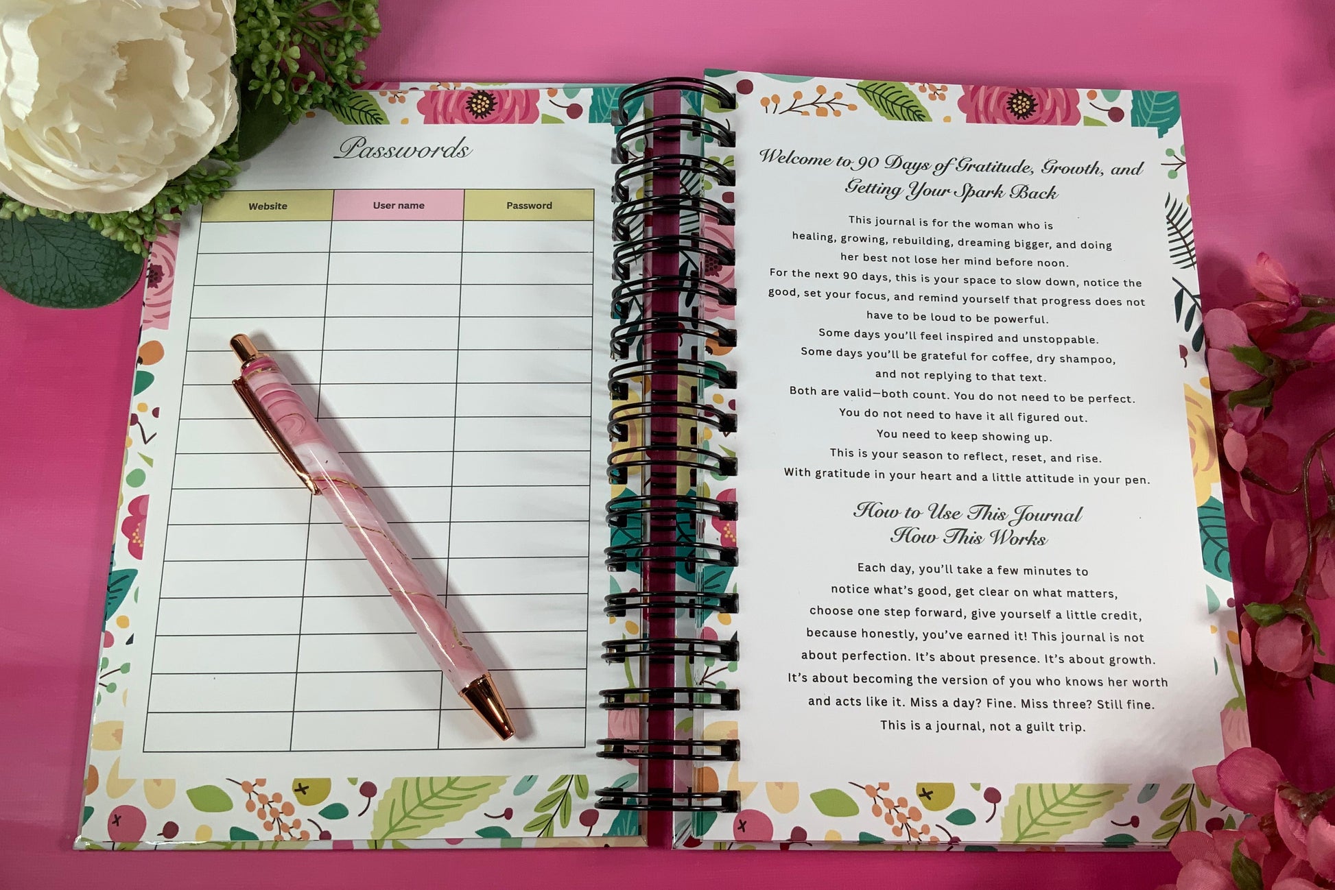 Open planner with floral design on a pink background with flowers and a pen.