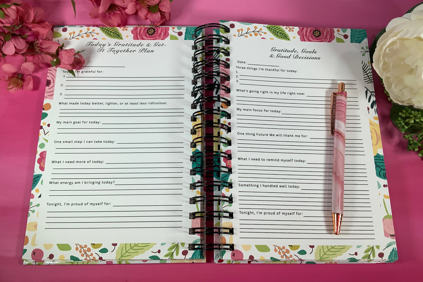 Open planner with floral design on a pink background with flowers and a pen.