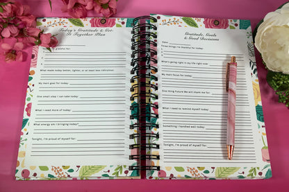 Open planner with floral design on a pink background with flowers and a pen.