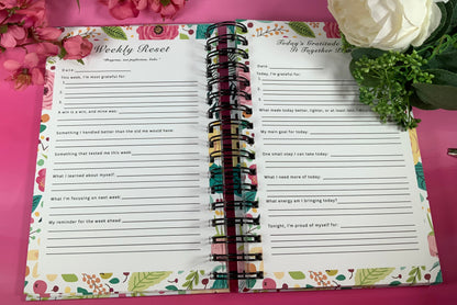 Open planner with weekly reset and reflection pages on a pink background with flowers.