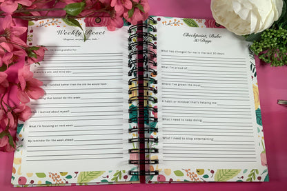 Open planner with weekly reset and gratitude lists on a pink surface with flowers.