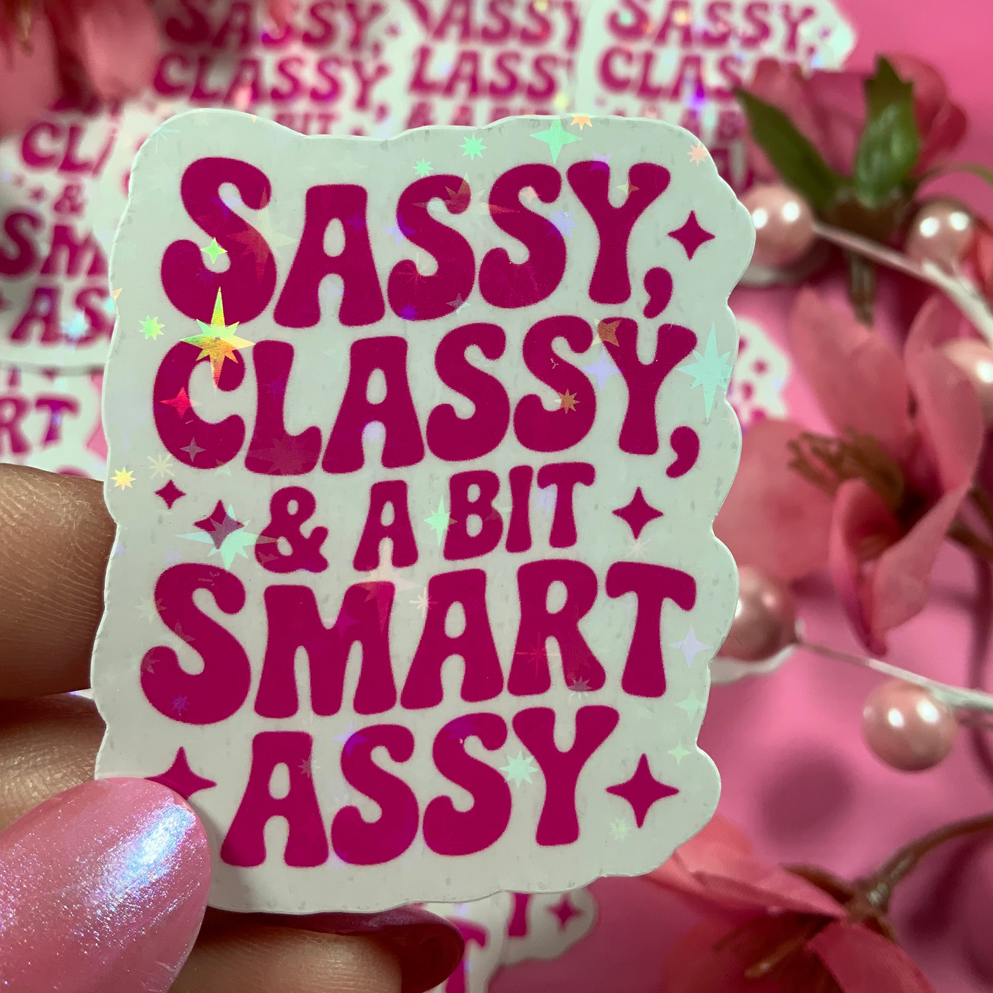 Sticker with 'Sassy, Classy, & A Bit Smart Assy' text held in front of pink flowers.