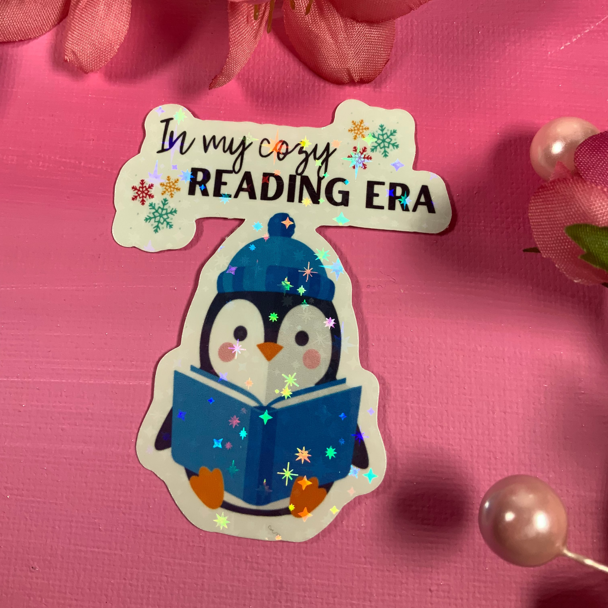 Sticker of a penguin reading a book with text 'In my cozy READING ERA' on a pink background with flowers.