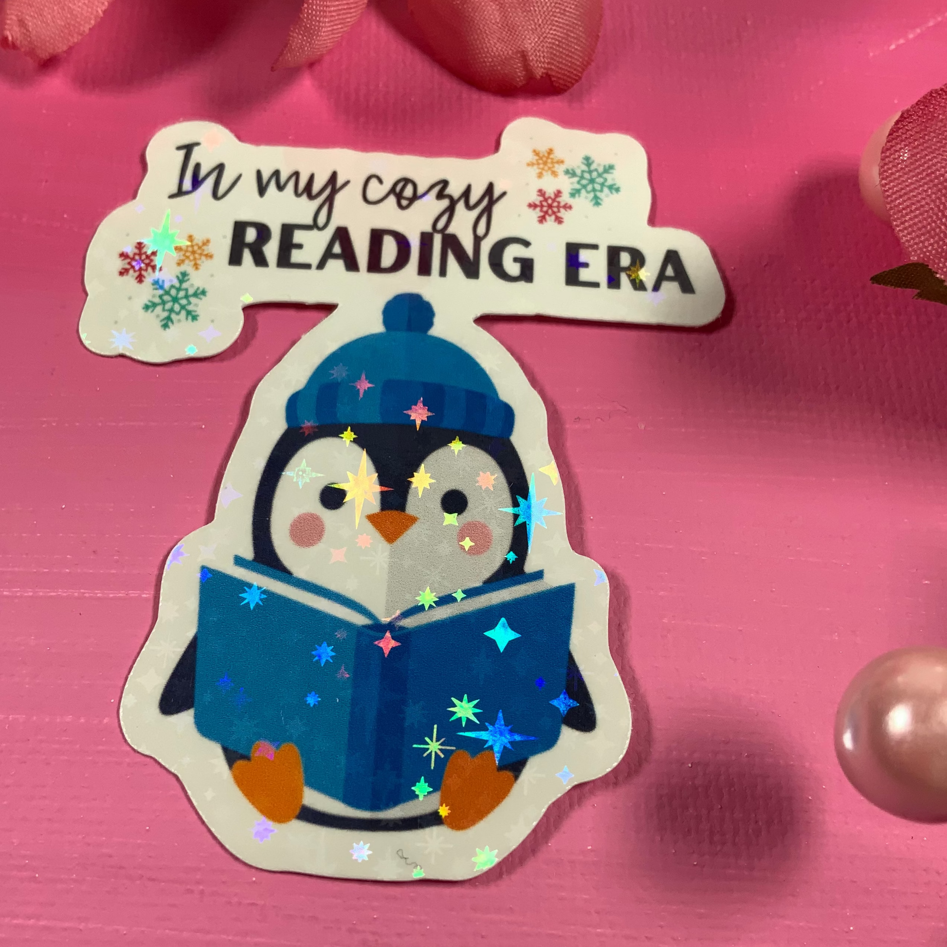 Sticker of a penguin reading a book with text 'In my cozy READING ERA' on a pink background.