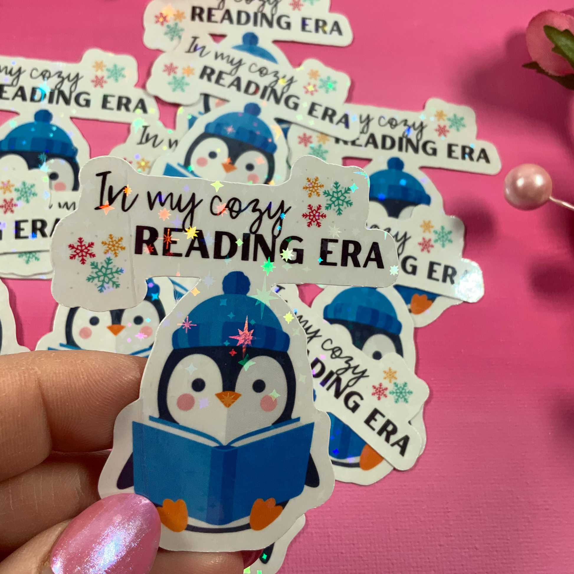 Sticker with penguin design and text 'In my cozy READING ERA' held against a pink background with flowers.