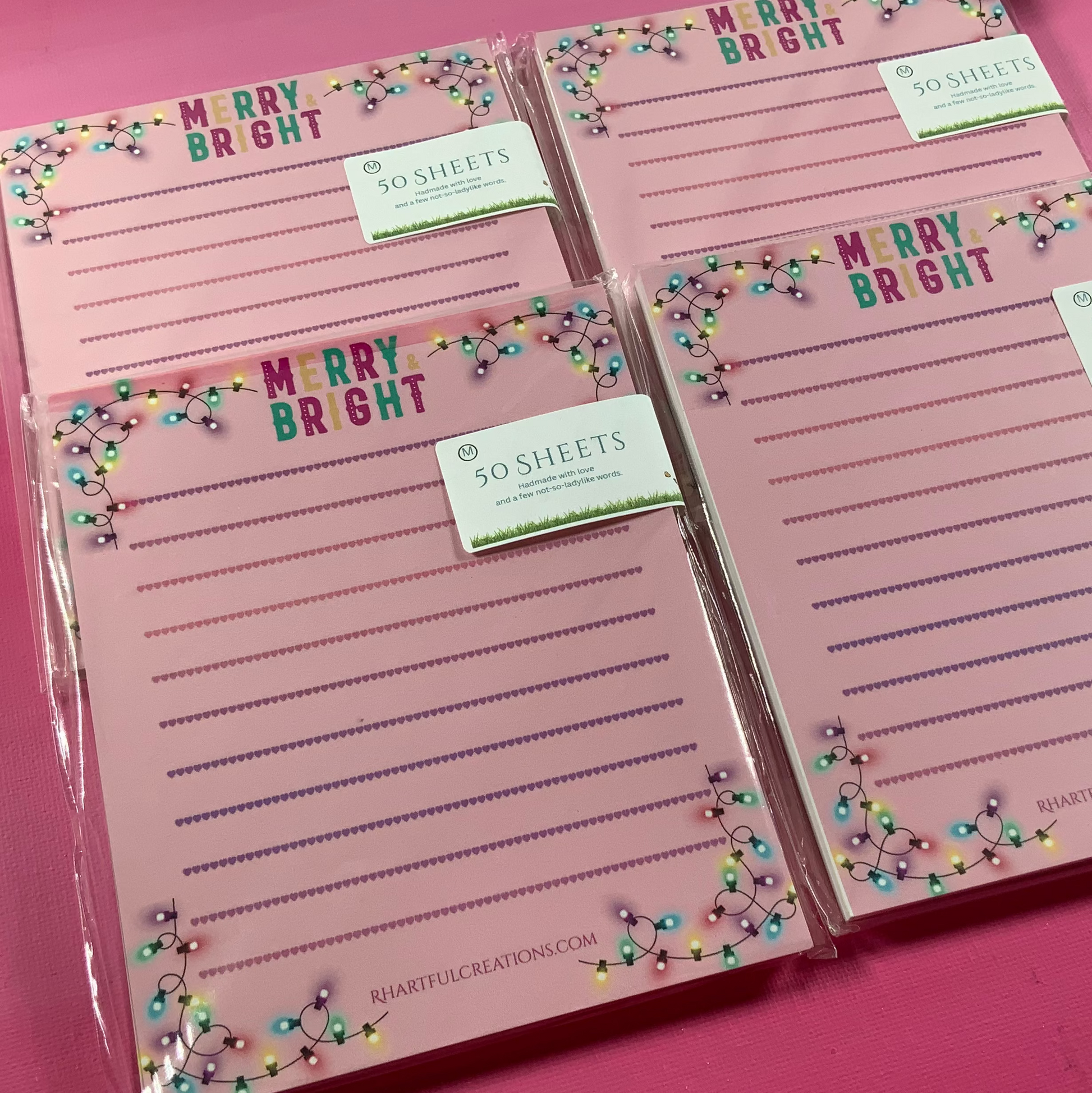 Two pink notepads with 'Merry Bright' text and decorative lights on a pink background.