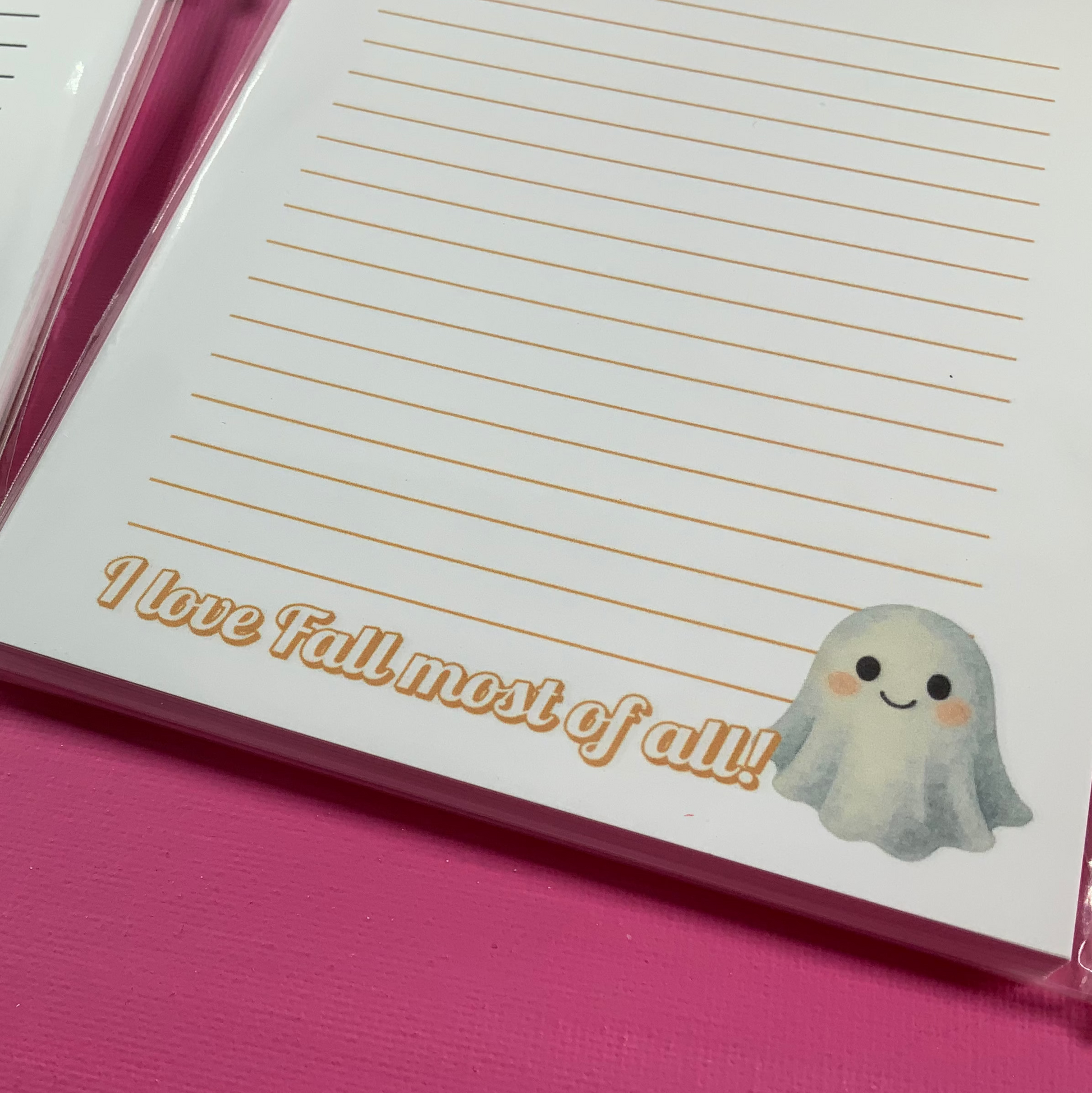 Two Halloween-themed notepads with ghost and pumpkin designs on a pink surface.
