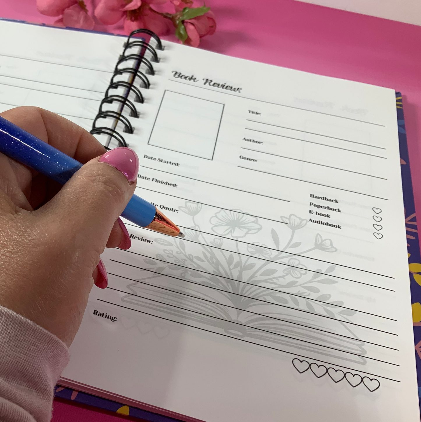 Hand holding a blue pen over a spiral-bound notebook on a pink surface with flowers.