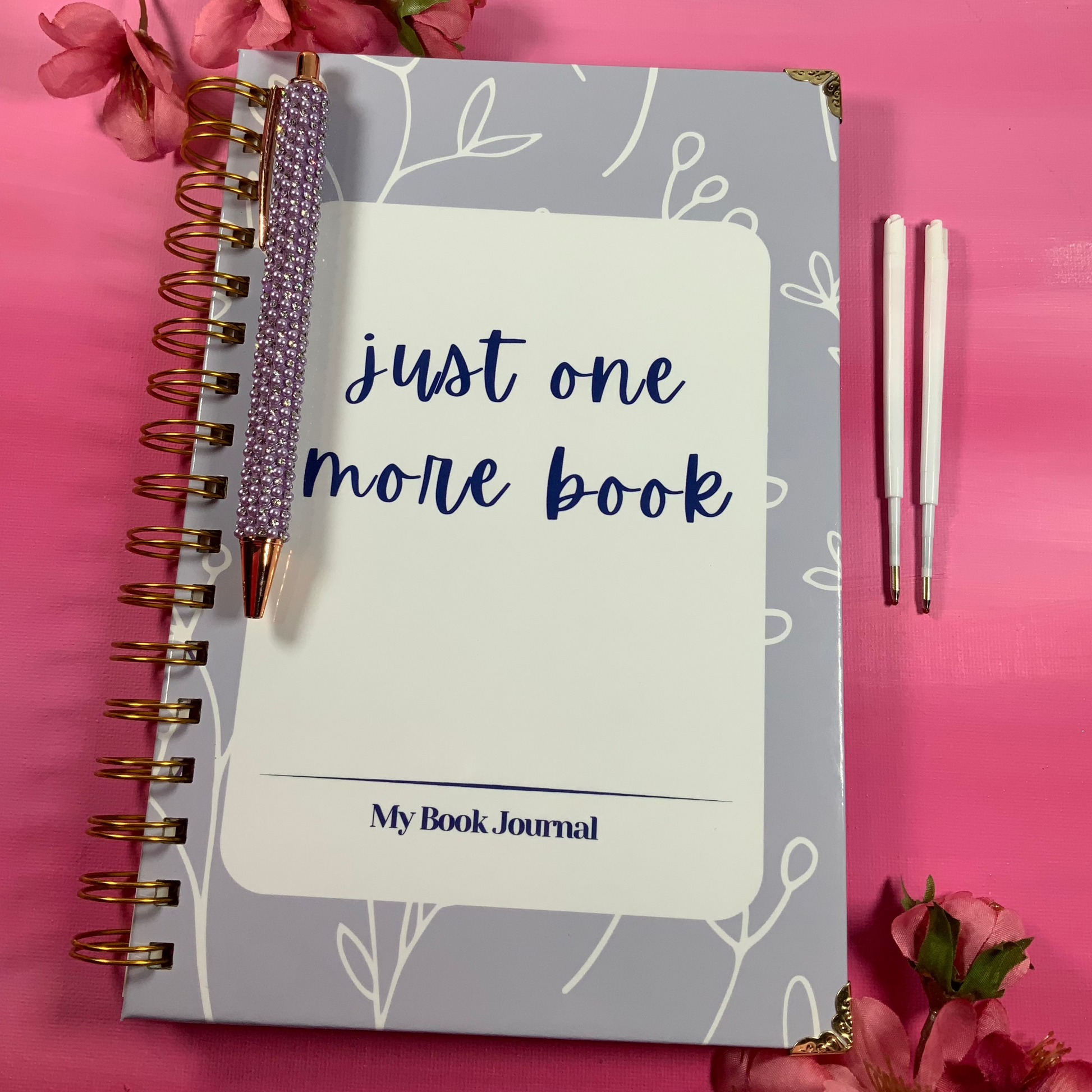 Notebook titled 'Just One More Book' with a pen on a pink background