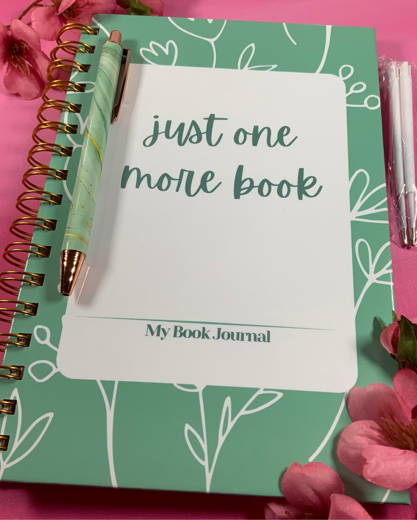 Green notebook titled 'Just One More Book' with a pen on a pink background