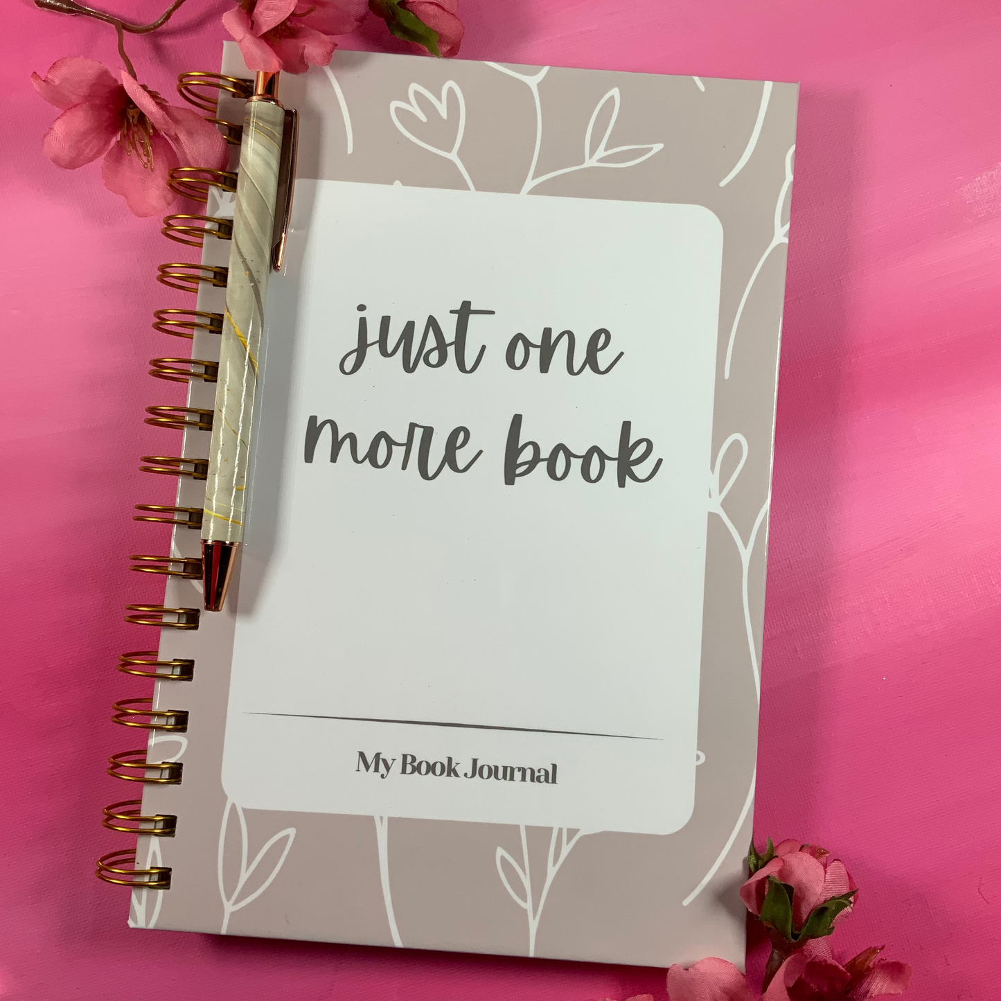 Notebook titled 'just one more book' with a pen on a pink background