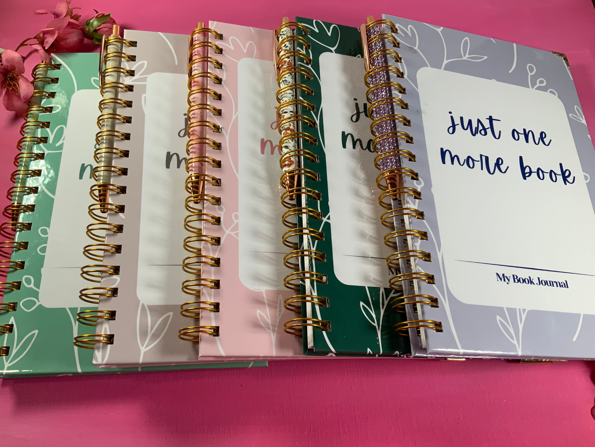 Three spiral-bound notebooks with different cover designs on a pink background.