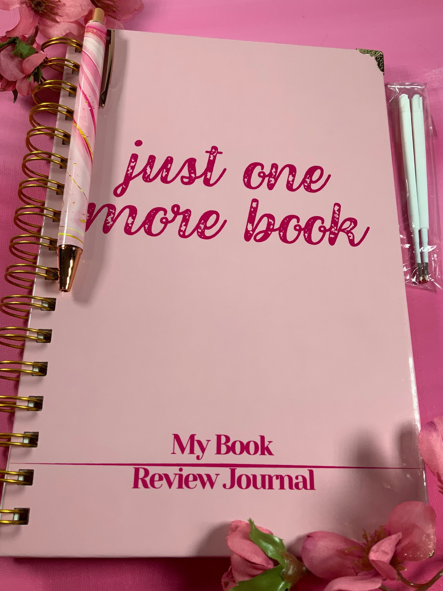Pink notebook titled 'just one more book My Book Review Journal' on a pink background with flowers.