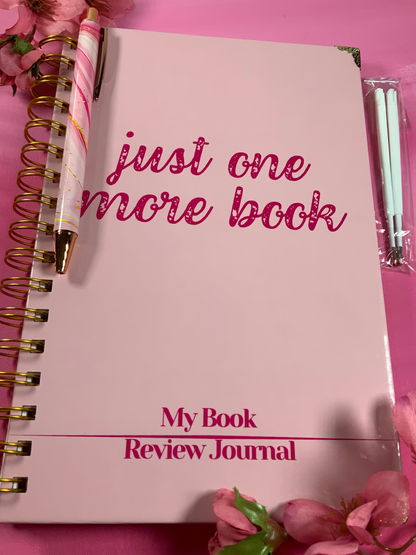 Pink notebook titled 'just one more book My Book Review Journal' on a pink background with flowers.