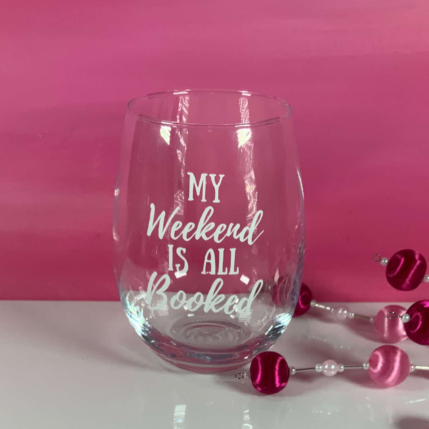 Clear glass with 'My Weekend is All Booked' text, pink beads on a white surface with pink background