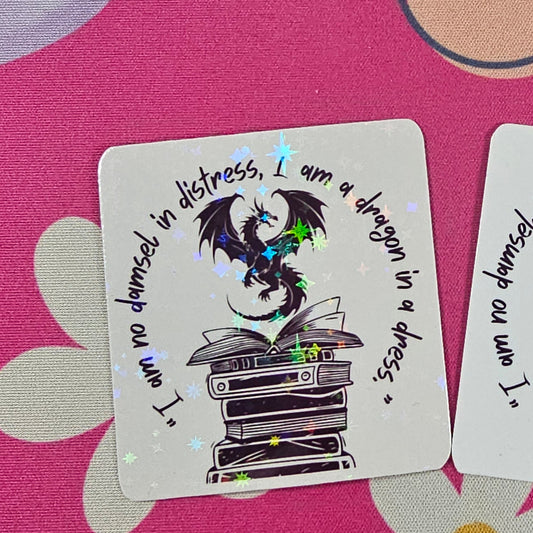 “I am no damsel in distress, I am a dragon in a dress” sticker featuring a black dragon sitting atop stacked books handmade by RH Artful Creations.