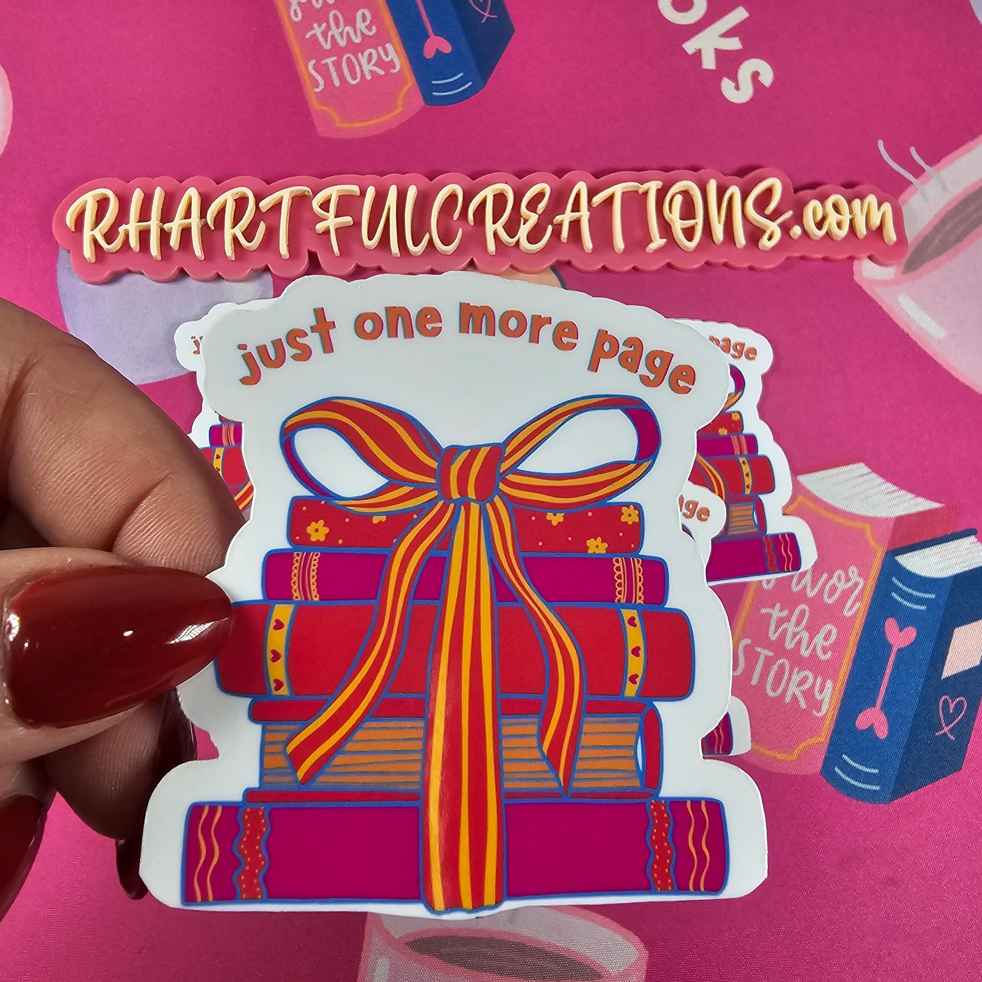 Hand holding “Just One More Page” sticker featuring a colorful stack of books tied with a ribbon, surrounded by cozy book-lover decor from RH Artful Creations.