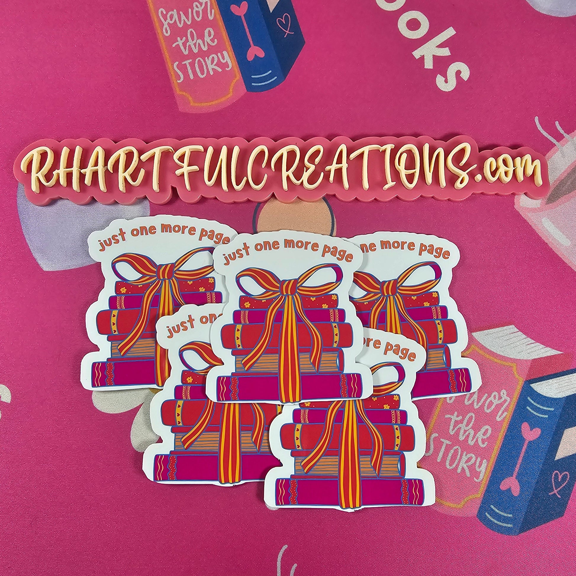 Hand holding “Just One More Page” sticker featuring a colorful stack of books tied with a ribbon, surrounded by cozy book-lover decor from RH Artful Creations.