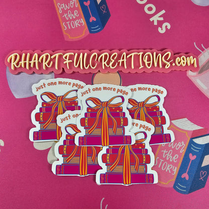 Hand holding “Just One More Page” sticker featuring a colorful stack of books tied with a ribbon, surrounded by cozy book-lover decor from RH Artful Creations.