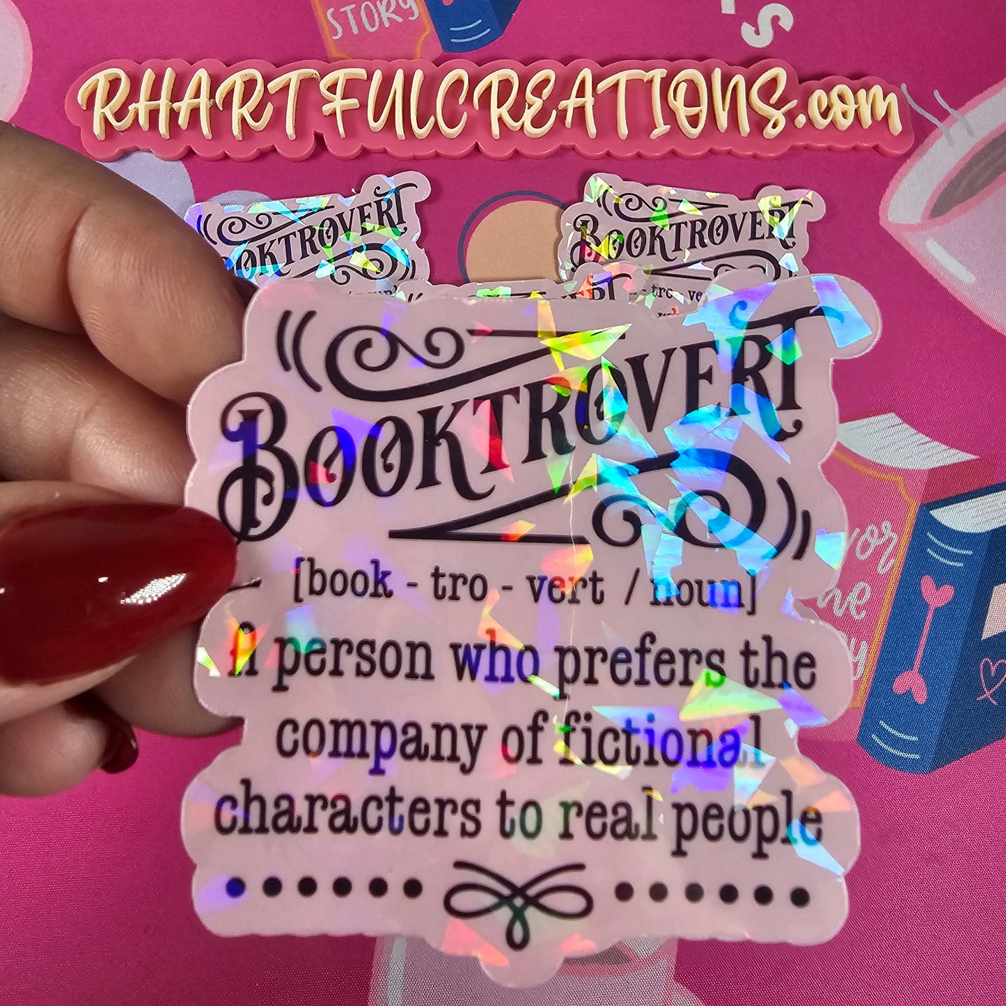 Holographic 'Booktrovert' sticker held by a hand with red nail polish against a pink background with text and graphics.