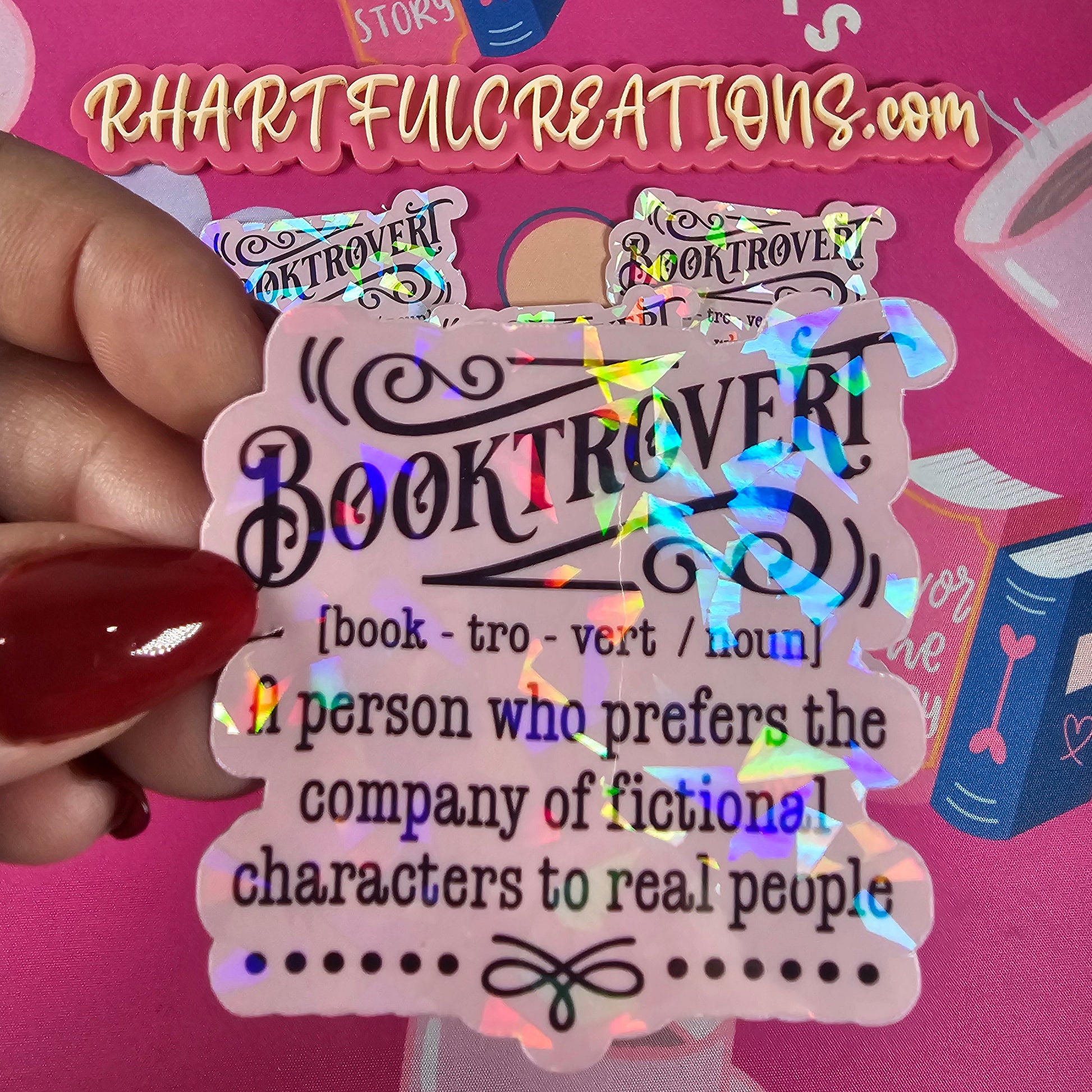 Holographic 'Booktrovert' sticker held by a hand with red nail polish against a pink background with text and graphics.