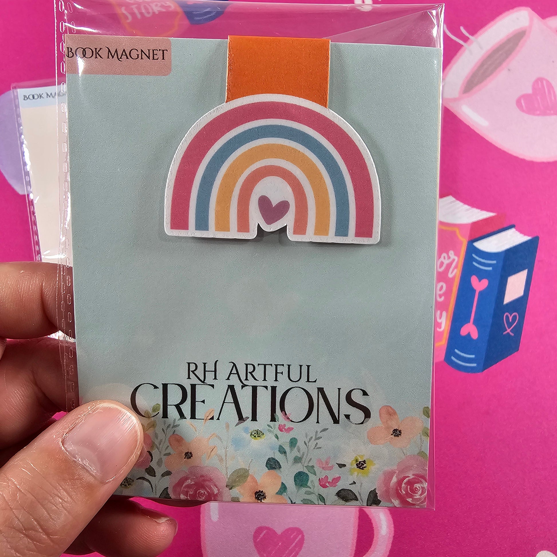 Hand holding a card with a rainbow design and 'RH Artful Creations' text against a pink background.