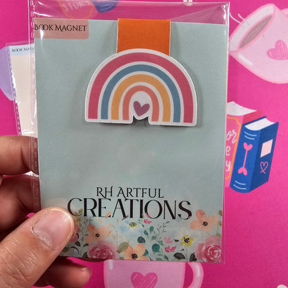 Hand holding a card with a rainbow design and 'RH Artful Creations' text against a pink background.