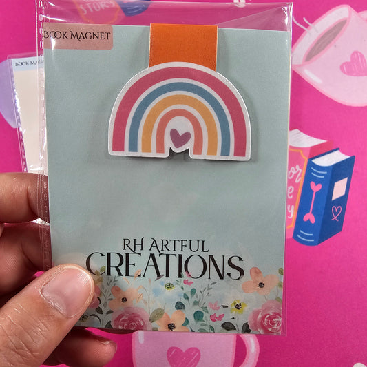 Hand holding a card with a rainbow design and 'RH Artful Creations' text against a pink background.