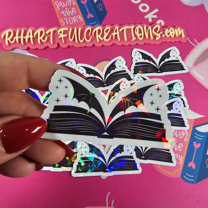 Holographic sticker shaped like an open book with magical wings, handmade by RH Artful Creations.