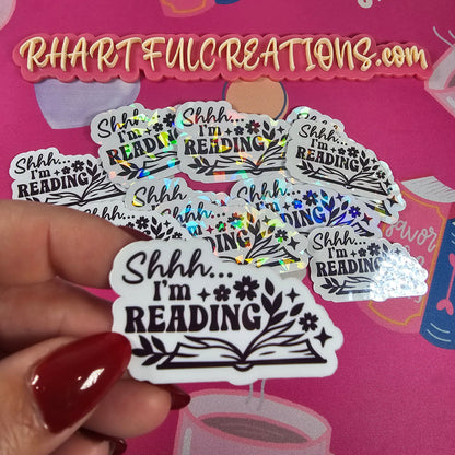 Hand holding multiple 'Shhh I'm Reading' stickers with a pink background featuring books and text.
